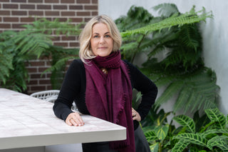 A woman with blonde hair wearing a black long-sleeve top and a deep merlot cashmere scarf by Kasmiri, leaning on a white tiled table in an outdoor patio setting with green plants and a brick wall background.