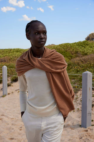 Person wearing a brown scarf and white outfit in a desert-like setting with greenery and blue sky.