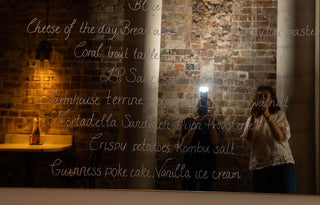 anna Kiousis and sarah garton taking a photo of a menu board with text on a brick wall.