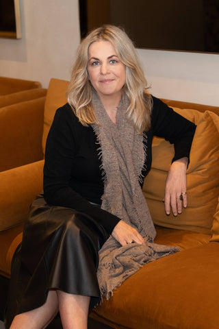 Samantha Theron sits on a mustard yellow couch wearing a black outfit and a Kasmiri Organic shawl wrap.