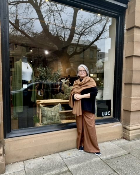 Lucy Given outside her luxury store, LUC DESIGN. LUCY wears KASMIRI cashmere travel wrap in spice and shows a cashmere travel wrap in olive green on a mannequin in the shop window